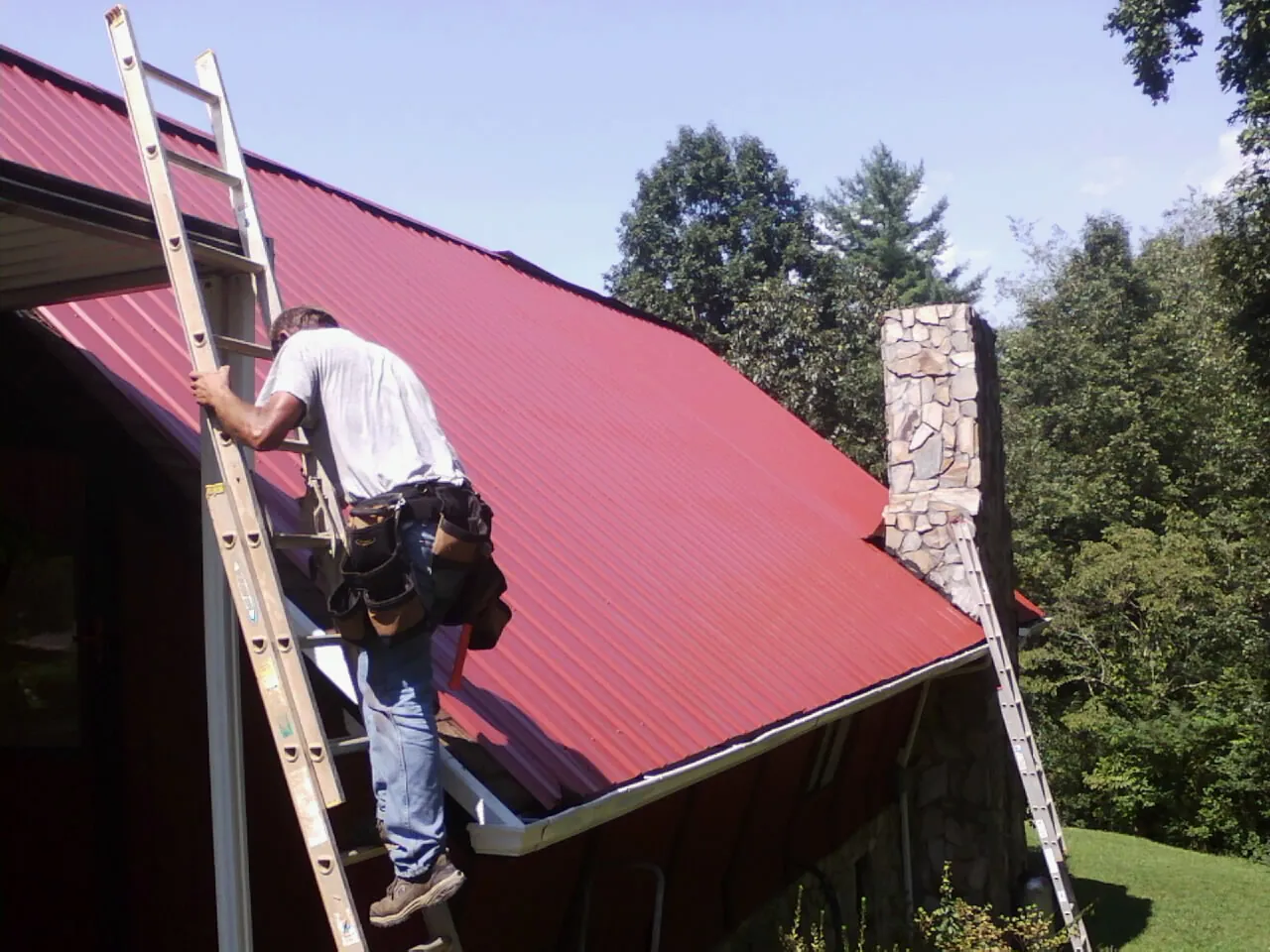 Roofer on a ladder beside a red metal roof and stone chimney — Soffit & Fascia Repair by Roofing Brothers in Wallisville